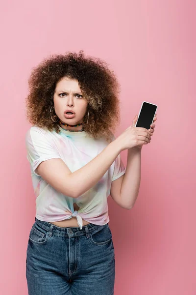 ピンクに隔離された巻き毛保持スマートフォンを持つ混乱した女性 — ストック写真