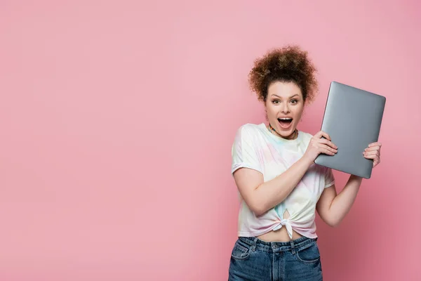 惊讶的卷曲女人拿着用粉色隔离的笔记本电脑 — 图库照片