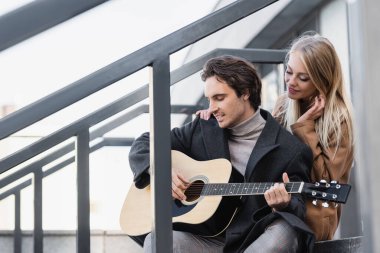Akustik gitar çalan, erkek arkadaşının omzuna dokunan mutlu sarışın kadın. 