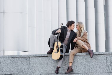 Sonbahar kıyafetli bir kadın korkulukta gitar çalan bir adamın yanında oturuyor.
