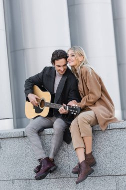 Güz kıyafetleri içinde neşeli bir kadın korkulukta oturup akustik gitar çalan bir adamın yanında oturuyor.
