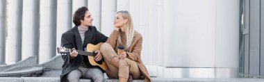 Hazır içkiyle mutlu bir kadın, açık havada akustik gitar çalan genç adama bakıyor.