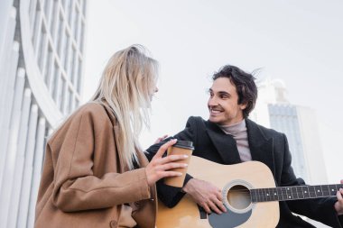 Açık havada gitar çalarken elinde içkiyle sarışın kadına bakan neşeli bir adam.