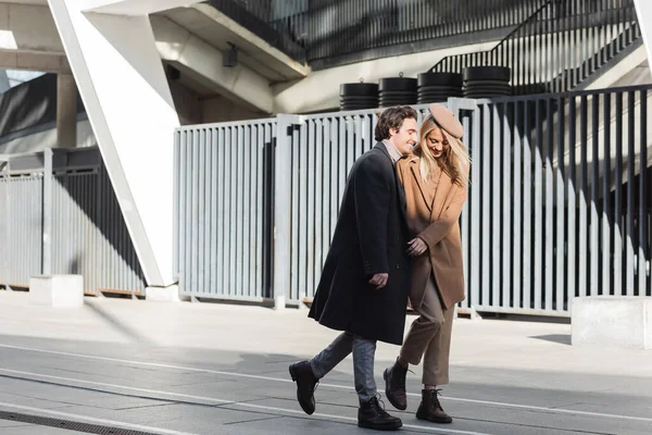 full length of stylish and happy couple in coats walking on urban street