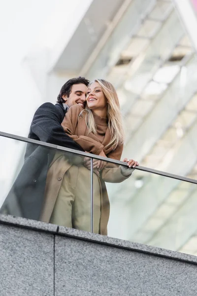 微笑的男人在玻璃栅栏边拥抱着快乐而时尚的女人的低视角 — 图库照片