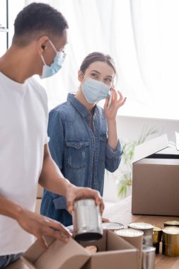 Tıbbi maskeli çok ırklı satıcılar konserve yiyecekler paketliyor ve online internet mağazasında akıllı telefondan konuşuyorlar. 