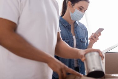 Bulanık Afrika kökenli Amerikalı meslektaşının yanında akıllı telefon kullanarak internet sitesinde konserve yiyecek paketleyen tıbbi maske satıcısı. 