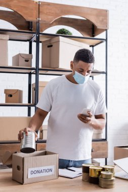Tıbbi maskeli Afrikalı Amerikalı satıcı elinde akıllı telefon ve bağış kutusunun yanında konserve yiyecek tutuyor. 