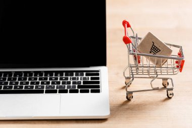 Toy shopping cart with package near laptop with blank screen on table  clipart