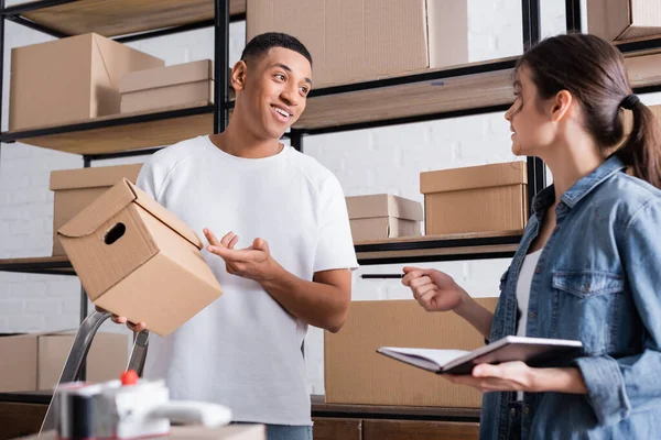 African american seller pointing at cardboard box near colleague with notebook in online web store