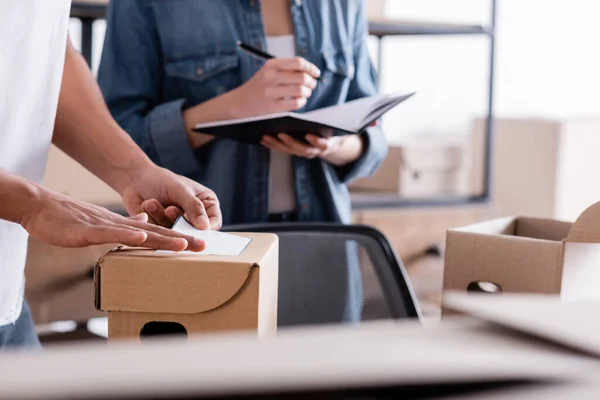 Cropped view of african american seller applying shipping label on package near colleague with notebook in online web store