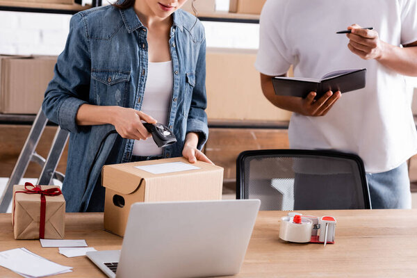 Обрезанный просмотр пакета сканирования продавца рядом с ноутбуком и африканским американским коллегой с ноутбуком в интернет-магазине 