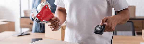 Cropped view of african american online web store owner holding scanner and adhesive tape near carton box, banner 