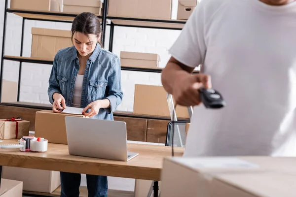 Seller fastening shipping label on cardboard box near laptop and african american colleague with scanner in online web store