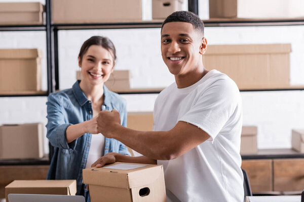 Cheerful interracial sellers doing fists bump near carton boxes in online web store 