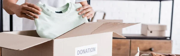 Cropped view of african american seller putting clothes in box with donation lettering, banner