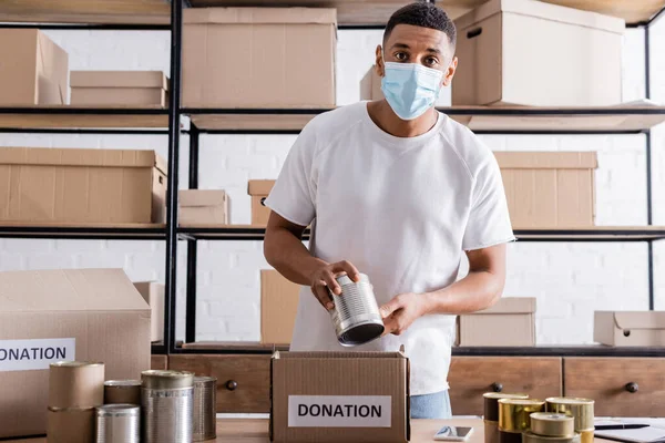 African american seller in medical mask holding can near donation box in online web store