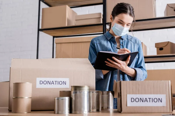 Seller in medical mask writing on notebook near canned food and boxes with donation lettering in online web store