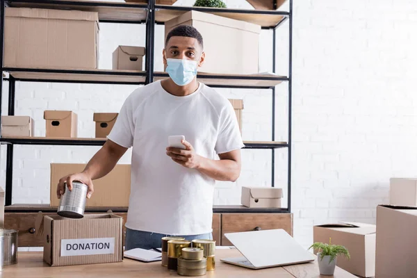 African american seller in medical mask holding smartphone near canned food and donation box in online web store