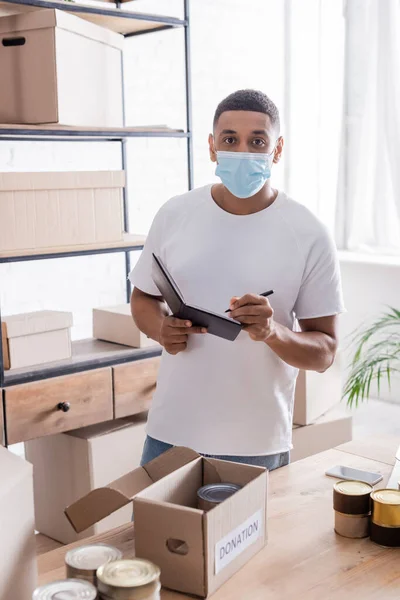 African american seller in medical mask holding notebook near cans and donation box in online web store