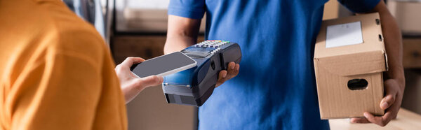 Cropped view of proprietor paying with smartphone near african american courier in online web store, banner 
