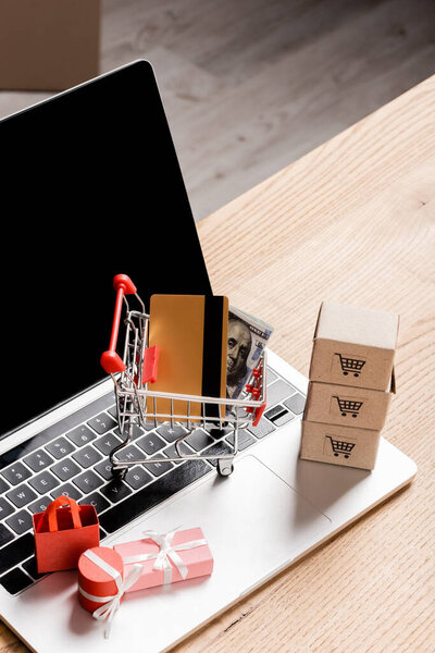 Toy shopping cart with credit card and money near packages on laptop with blank screen 