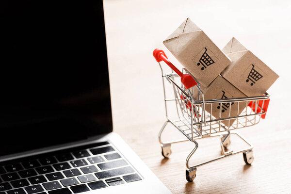 Packages in toy shopping cart near laptop with blank screen on table 