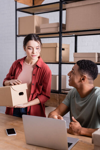 Улыбающийся африканский американский продавец смотрит на коллегу с пакетом рядом с устройствами в интернет-магазине 