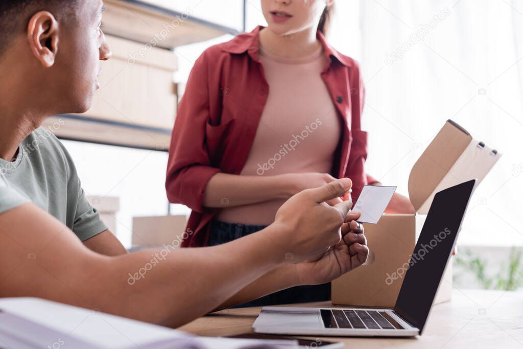 African american seller holding shipping label near laptop and colleague with carton box in store 