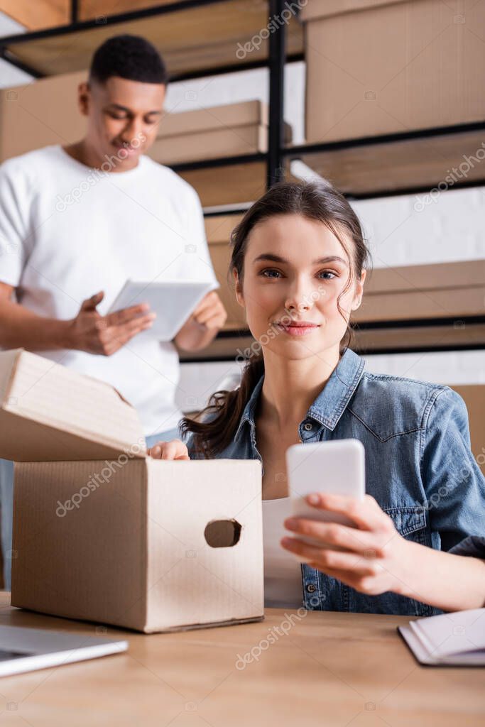 Young seller holding blurred smartphone near carton box and blurred african american colleague in store 