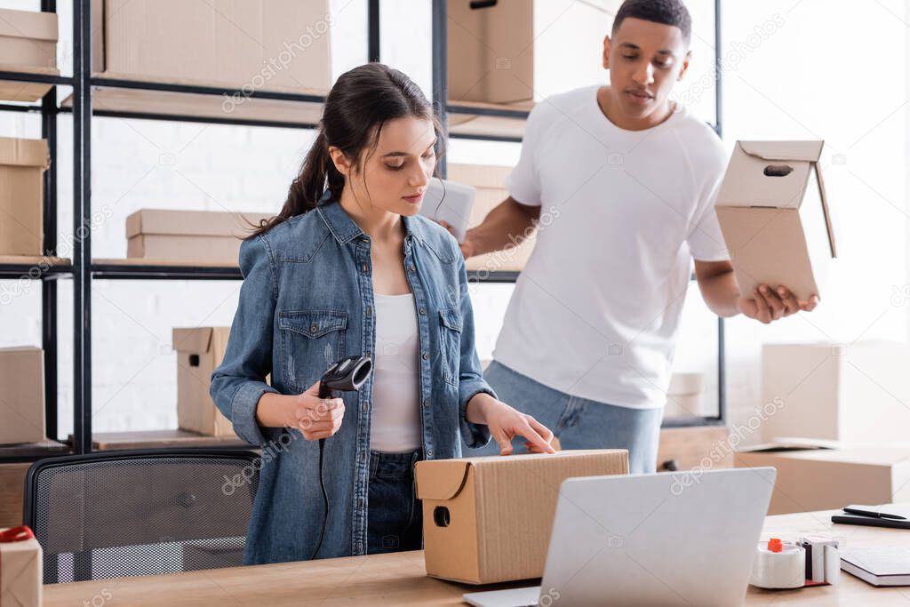 Seller holding scanner near carton box, laptop and colleague in online web store 