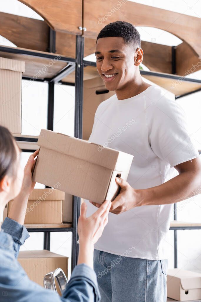 Smiling african american seller holding carton box with blurred colleague in online web store 