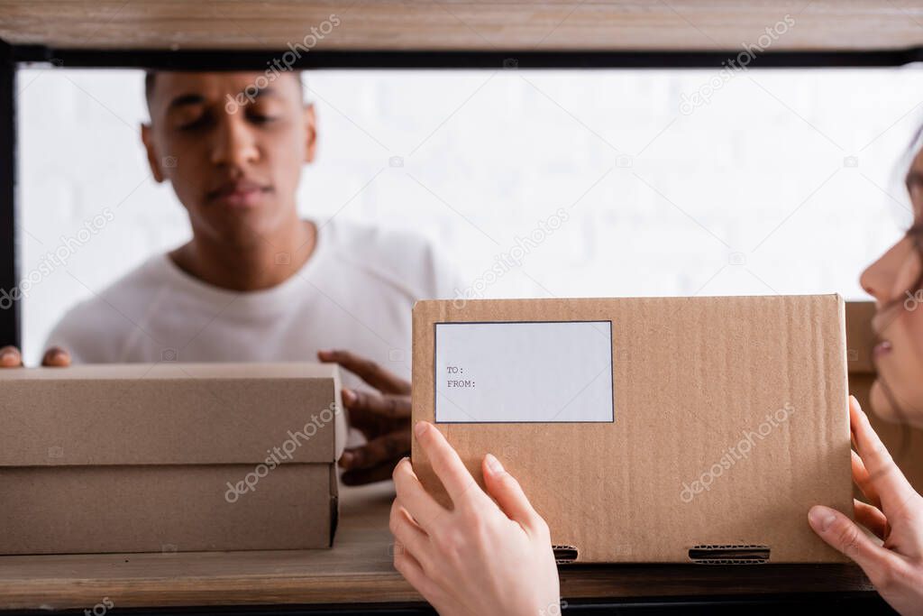 Blurred seller holding cardboard box with shipping label near blurred african american colleague in online web store 