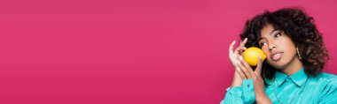 Kıvırcık Afrikalı Amerikalı kadın limon tutuyor ve pembe pankartta izole bir şekilde yüzünü çeviriyor.