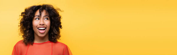 astonished african american woman with curly hair looking aside isolated on yellow, banner