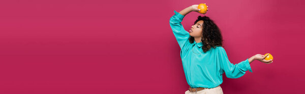 стильная африканская американка в голубой блузке позирует с апельсинами изолированы на розовый, баннер