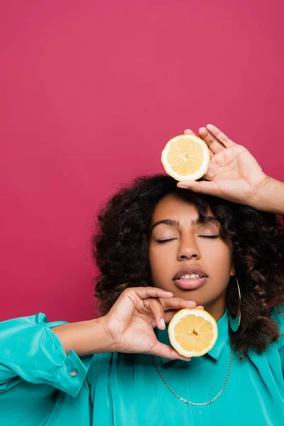 african american woman with closed eyes holding halves of juicy lemon isolated on pink