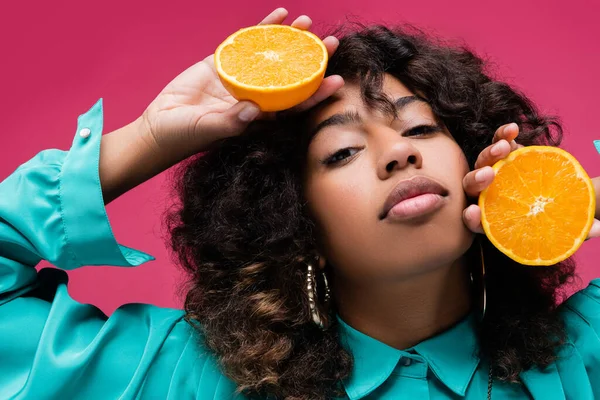 african american woman with wavy hair holding halves of juicy orange isolated on pink