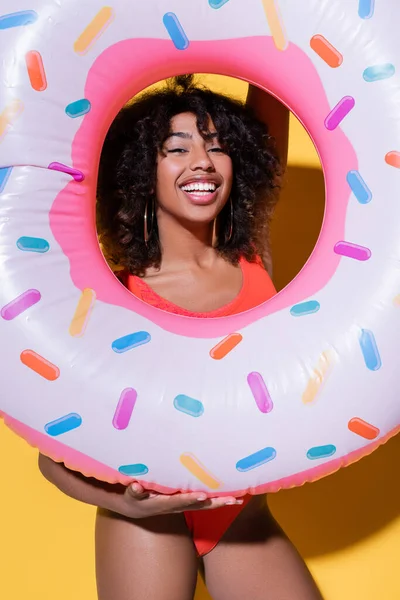 joyful african american woman with swim ring looking at camera on yellow background