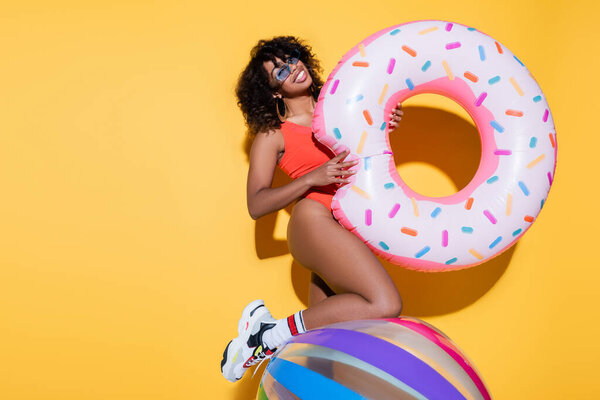 brunette african american woman in beachwear holding swim ring near inflatable ball on yellow background