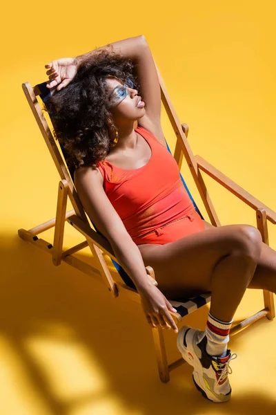 身穿墨镜和泳衣的黑发非洲裔美国女人躺在黄色背景的躺椅上 — 图库照片
