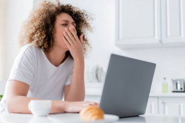 Uykulu kadın kahvaltı sırasında laptopun yanında esnerken ağzını eliyle kapatıyor.