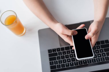 Kesilmiş kadının bilgisayarının yanında boş ekranı ve bir bardak portakal suyu olan akıllı telefonu kullanışının üst görüntüsü.