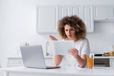 worried woman gesturing near digital tablet and laptop in kitchen clipart