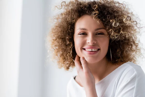 happy young woman with wavy hair touching face and smiling at camera