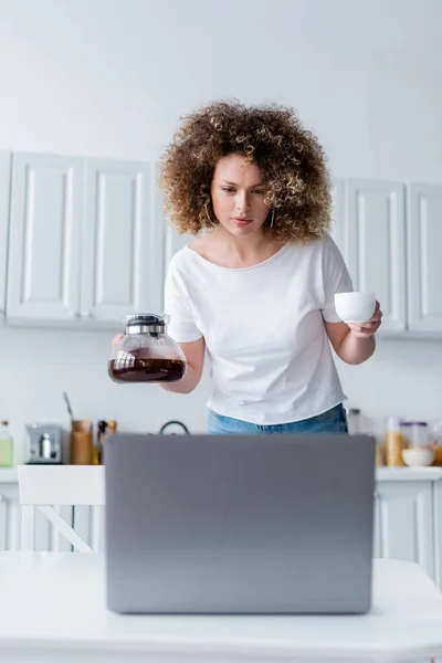 年轻女子一边拿着杯子和咖啡壶 一边看着厨房里的笔记本电脑 — 图库照片