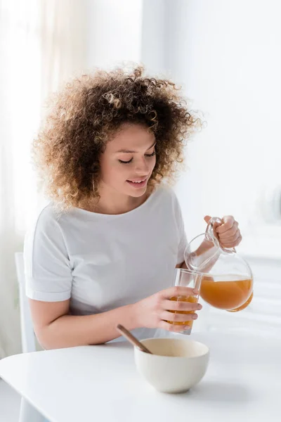 年轻而积极的女人 一头波浪形的头发 把橙汁从罐子倒进杯子里 — 图库照片