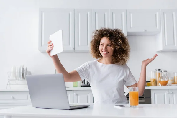 在厨房笔记本电脑旁的数字平板电脑上 一头波浪形头发的快乐女人正在自拍 — 图库照片
