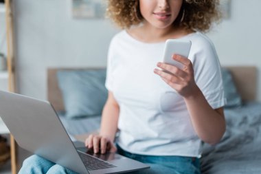 Yatak odasında dizüstü bilgisayarı ve cep telefonu olan gülümseyen kadının kısmi görüntüsü