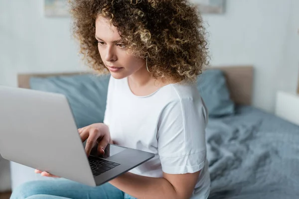 Młoda Kobieta Falowanymi Włosami Pomocą Laptopa Podczas Siedzenia Łóżku — Zdjęcie stockowe